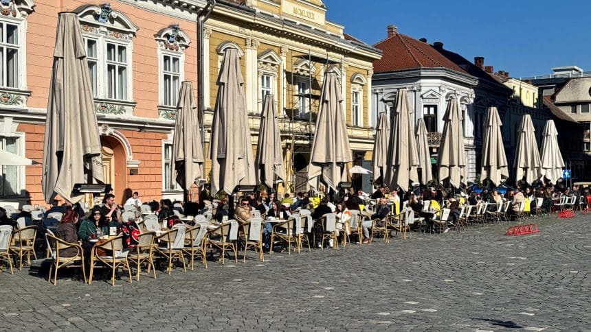 Oameni relaxându-se la mesele unei terase dintr-o piață istorică din Timișoara într-o zi însorită de primăvară