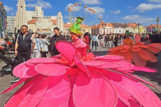 Studente costumate în flori defilând prin Piața Libertății din Timișoara în cadrul Paradei Florilor, însoțite de instrumentiști.