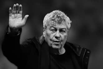 Fotografie alb-negru a lui Mircea Lucescu, zâmbind pe banca tehnică, reprezentând deceniile de glorie în fotbalul internațional.