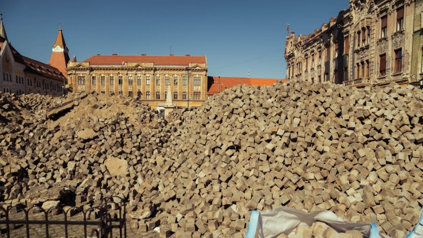 Muncitori sortând și curățând manual blocurile de piatră cubică scoase din pavajul Pieței Traian pentru a fi refolosite.