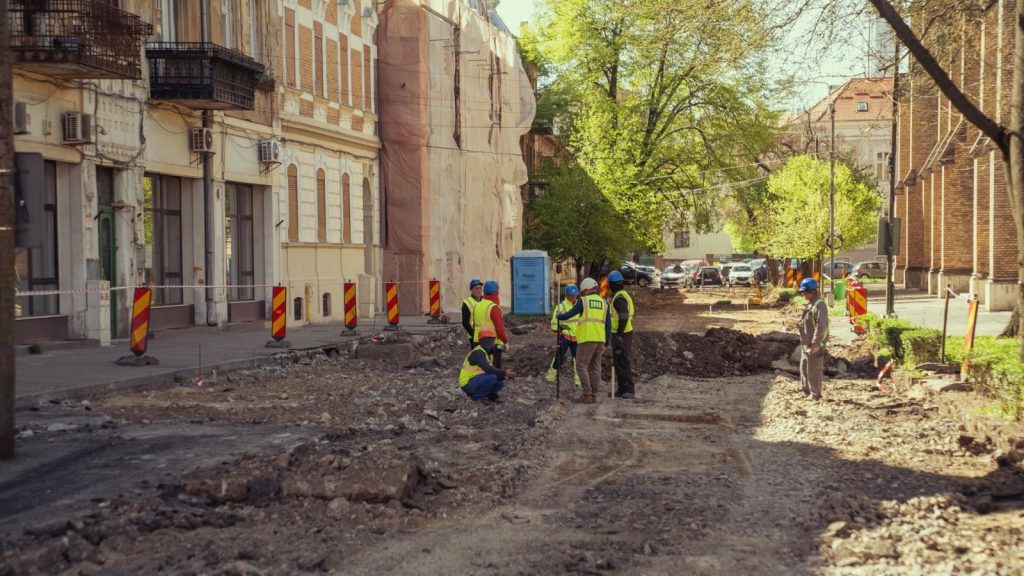 Muncitori sortând și curățând manual blocurile de piatră cubică scoase din pavajul Pieței Traian pentru a fi refolosite.