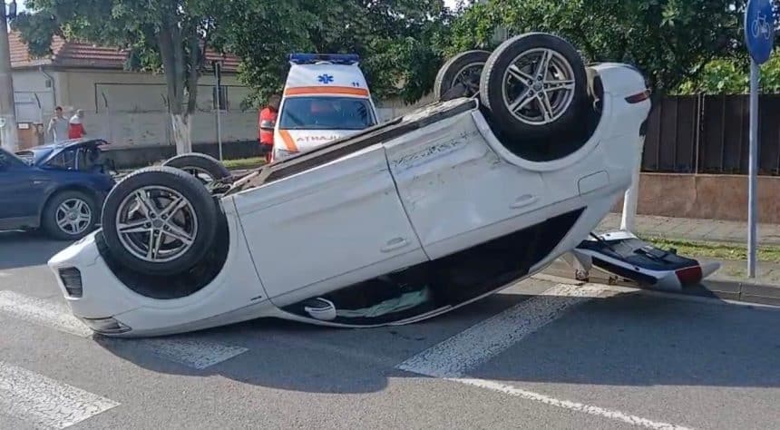 Un autoturism răsturnat pe plafon în mijlocul unei intersecții din Timișoara, înconjurat de echipaje de intervenție și bucăți de caroserie.