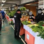 Agenți ai Poliției Locale Timișoara verificând atestatele de producător și cântarele comercianților la o tarabă de legume dintr-o piață agroalimentară.